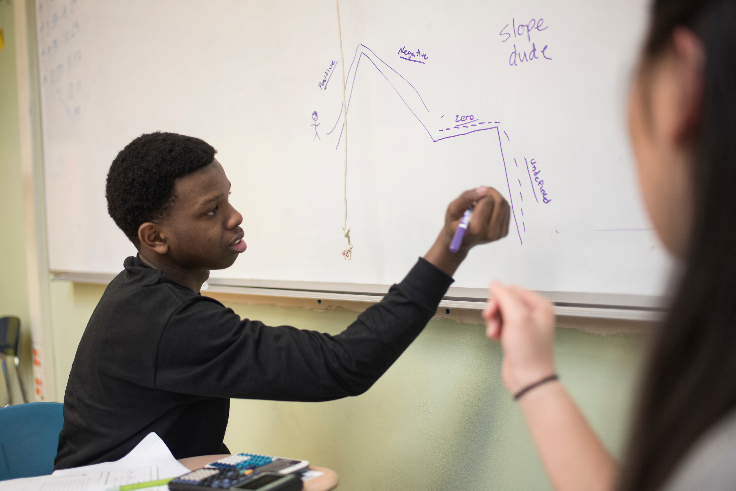 Male writing on whiteboard with female sitting beside him in meeting.