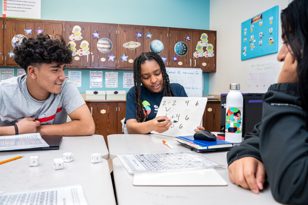 A group of students are working together at a table, with a tutor showing a math problem to the students.